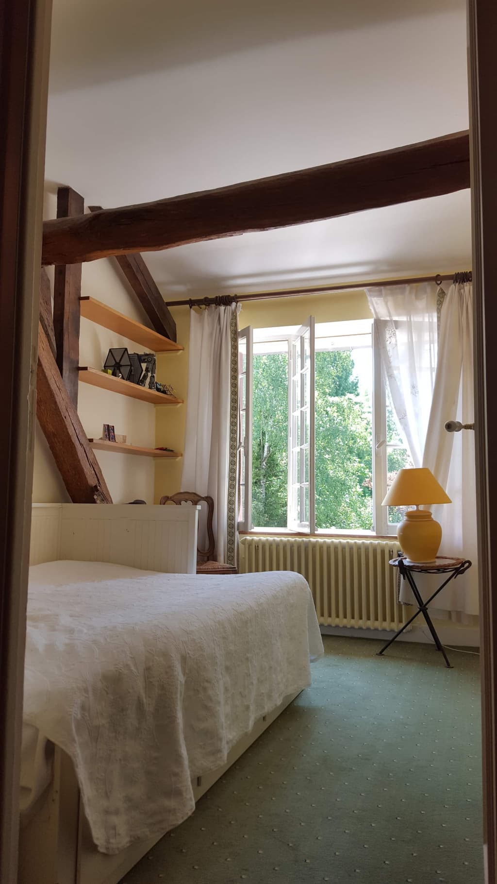 Zimmer unter der Dachschräge, Bett mit weißer Tagesdecke, offenes Fenster mit Blick auf die Bäume